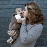 Loving mother kisses her baby, showcasing a tender family moment against a dark brick wall.