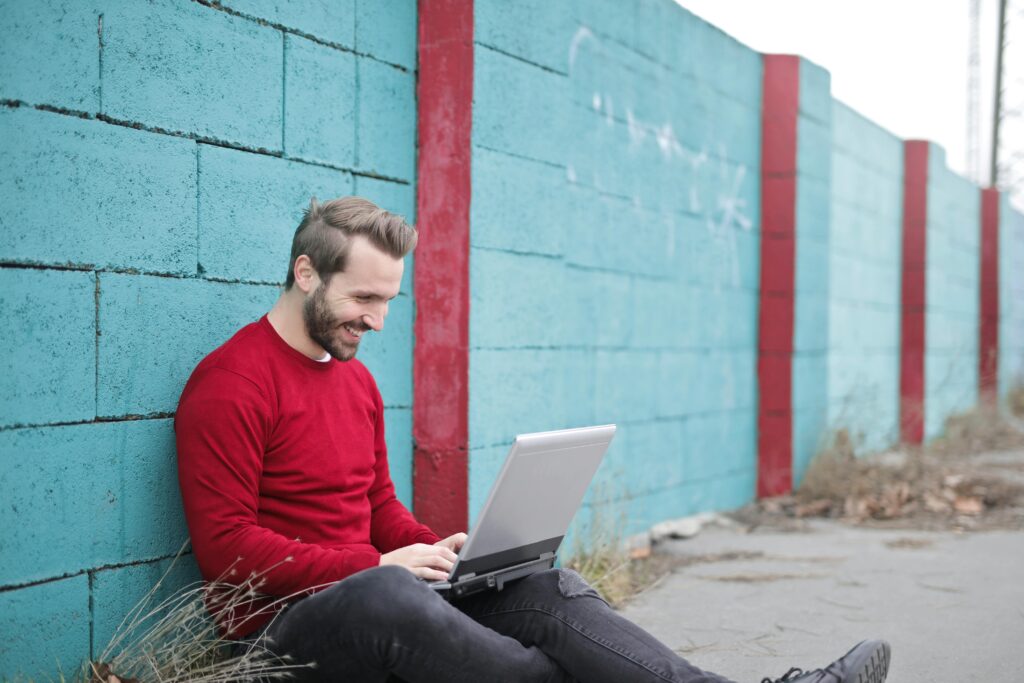 pexels photo 941572 941572 A man smiling and using a laptop outdoors against a vibrant blue wall, representing remote work.